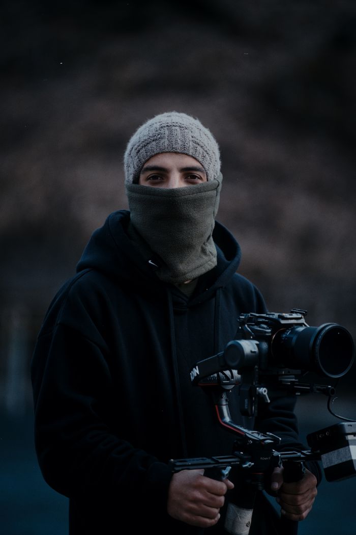 Portrait of a cameraman in a hoodie holding professional camera gear outdoors.